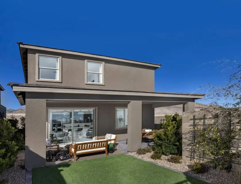A modern two-story house with beige stucco exterior, a covered patio with outdoor seating, large sliding glass doors, and neatly landscaped yard with artificial grass under a clear blue sky.