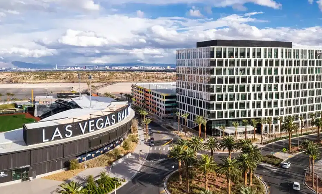 Aerial view of Las Vegas Ballpark and a modern office building, surrounded by palm trees and roads, with mountains and the Las Vegas skyline in the background under a partly cloudy sky.