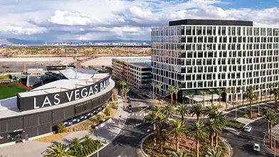 Aerial view of a modern Las Vegas street with palm trees, a large building labeled Las Vegas Ballpark, and a multi-story office building under a partly cloudy sky.