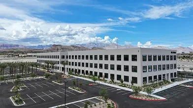 A large, modern, three-story office building with many windows sits in a nearly empty parking lot under a bright blue sky with scattered clouds and mountains in the background.