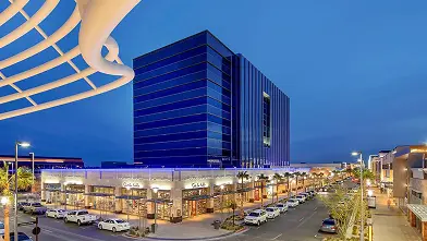 A modern cityscape at dusk features a tall glass building surrounded by well-lit shops, restaurants, parked cars, and a curved white architectural structure in the foreground.