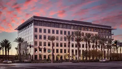 A modern, rectangular office building with large windows and surrounded by palm trees, set against a colorful sunset sky with pink and purple clouds.