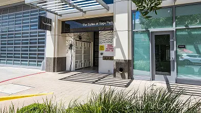 Entrance to a modern building with “The Sables at Sagemont” above the doorway, large windows, greenery in the foreground, and shadows cast on the sidewalk from a glass awning overhead.