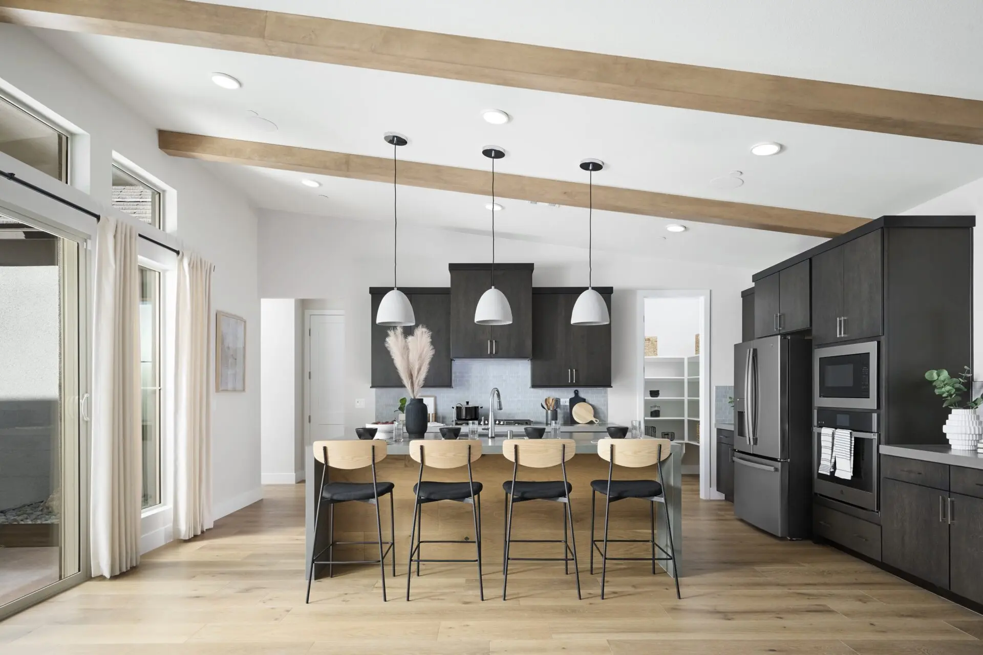 Modern kitchen with dark cabinets, stainless steel appliances, and a large island. Four wooden bar stools face the island, which is decorated with a vase of pampas grass. Light wood beams run across the ceiling.