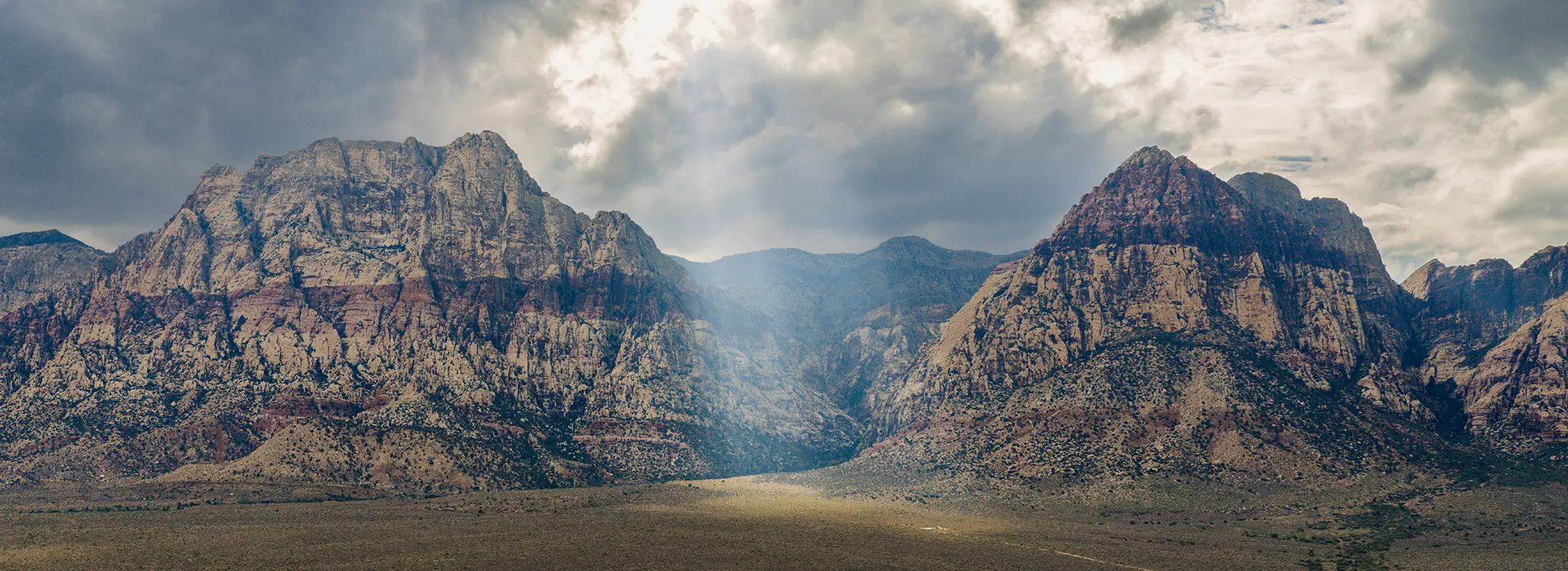 default Dramatic clouds hover above two rugged, rocky mountains with sunlight streaming through a gap, illuminating a patch of the arid landscape below.
