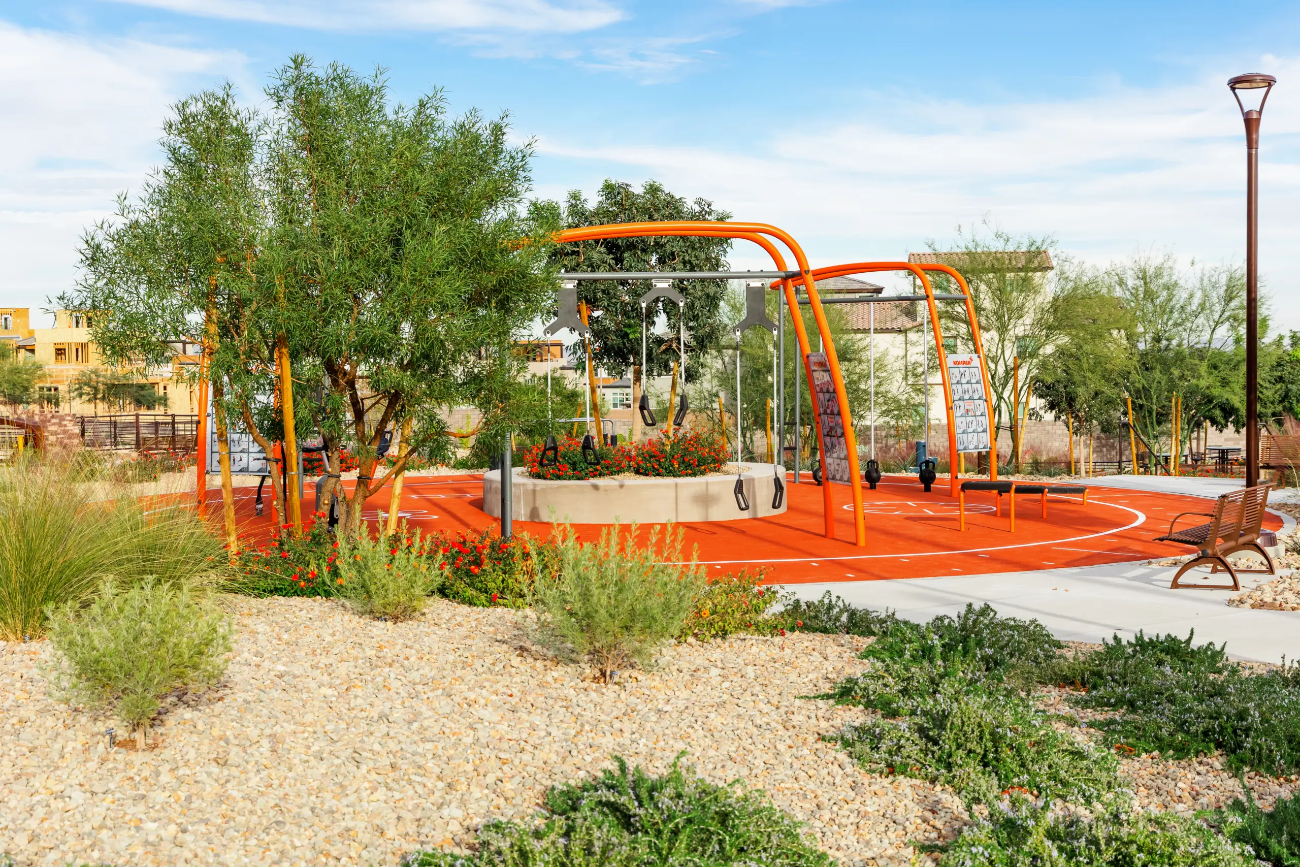 A modern playground with bright orange equipment, swings, and a soft rubber surface. The area is landscaped with rocks, shrubs, and trees, with benches and a lamppost nearby under a blue sky.