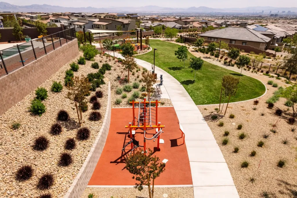 Aerial view of a suburban park with a red outdoor exercise area, landscaped plants, a walking path, green grass, and nearby houses under a hazy sky.