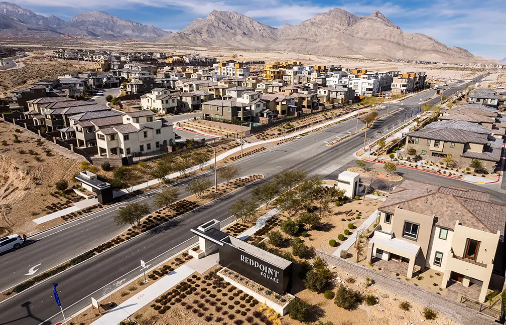 Redpointe Square Monument Aerial view of a suburban neighborhood with rows of modern houses, landscaped yards, and wide streets. A sign reading REDPOINT marks the entrance, with desert mountains and dry terrain in the background.