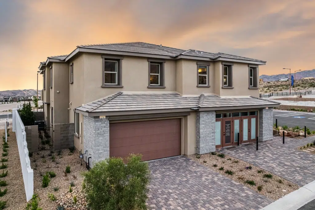 Two-story modern house with beige stucco and stone exterior, large windows, a double garage, and a paved driveway, set against a sunset sky and desert landscape.