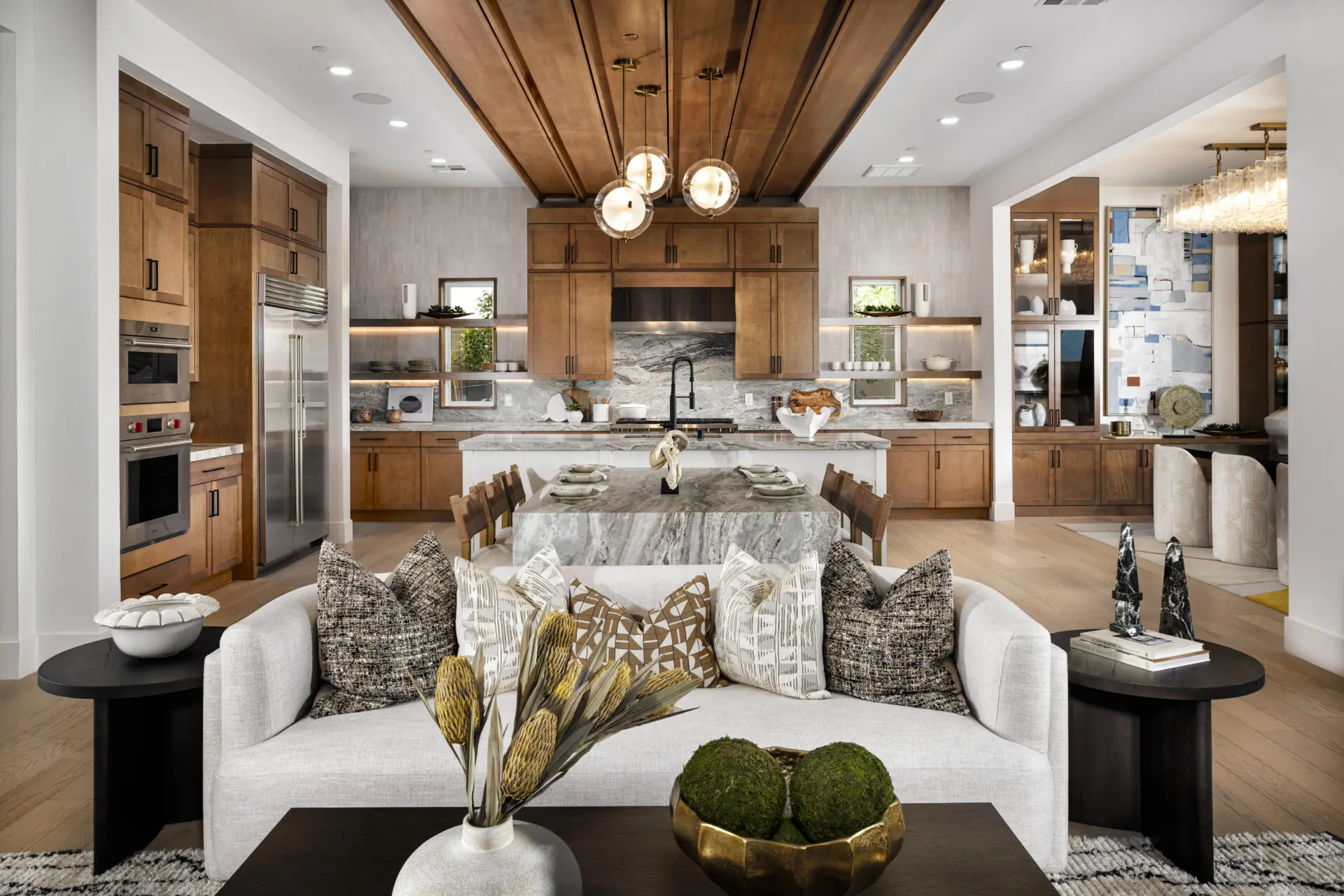 Modern open-concept kitchen and living area with wood cabinets, marble island, pendant lights, decorative pillows on a white sofa, and stylish accents on tables and shelves.