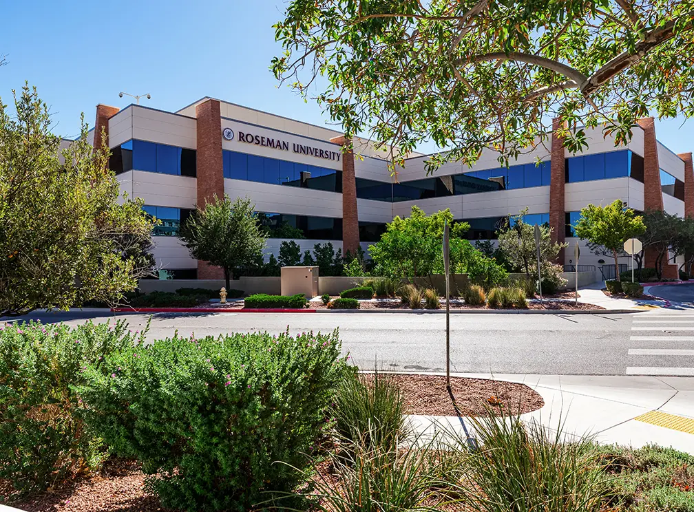 Summerlin Roseman University A modern, three-story building with Roseman University signage, large windows, and surrounded by trees and landscaping on a sunny day. A crosswalk and sidewalk are visible in the foreground.