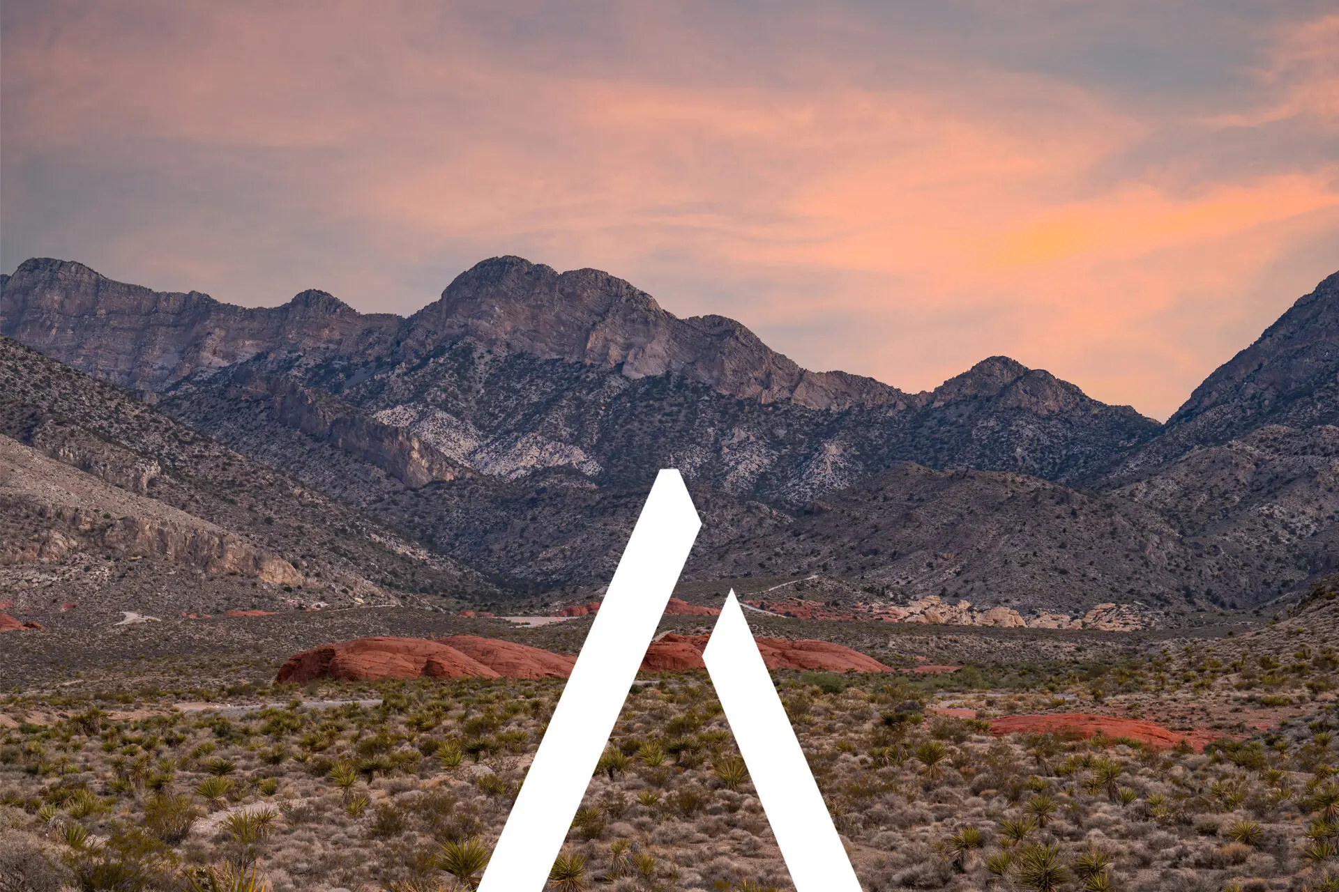 Desert landscape with rugged mountains in the background under a pink and orange sunset sky. In the foreground, there is a white angular graphic resembling an inverted V shape on the ground.
