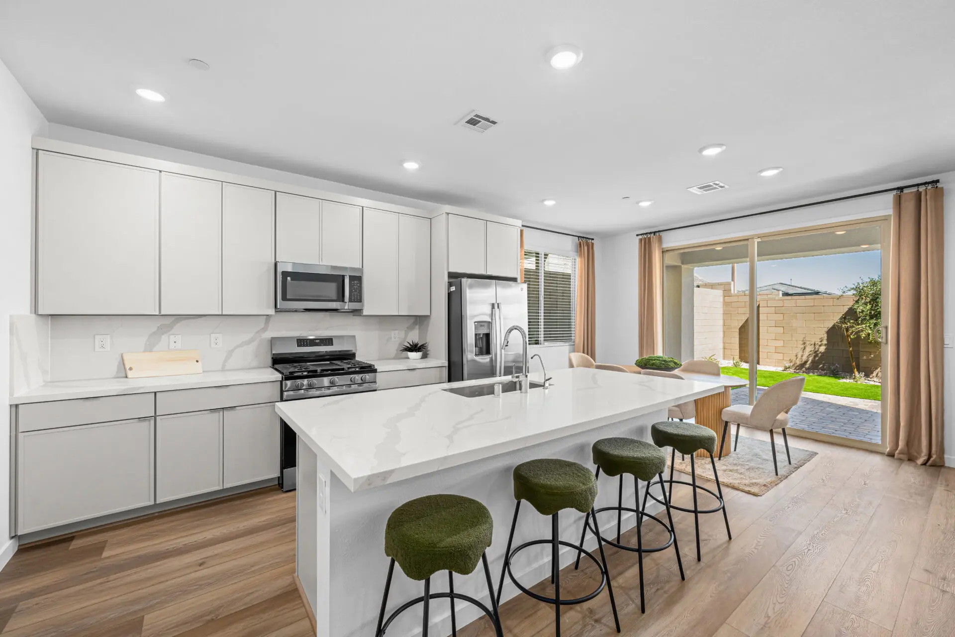 Modern kitchen with white cabinets, stainless steel appliances, marble island with sink, and four green cushioned bar stools. Dining area with table and chairs next to large sliding glass doors opening to a backyard.