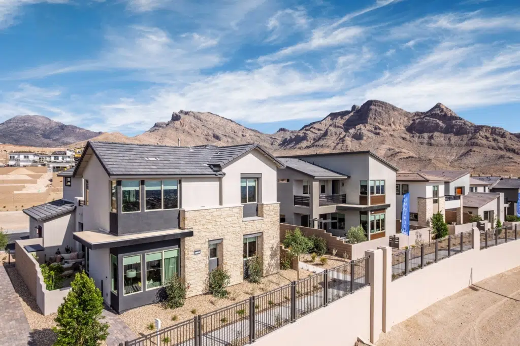 Modern two-story homes with flat roofs and large windows line a residential street in a desert area, with rocky mountains and blue sky in the background. A fence and landscaped yards are visible in the foreground.