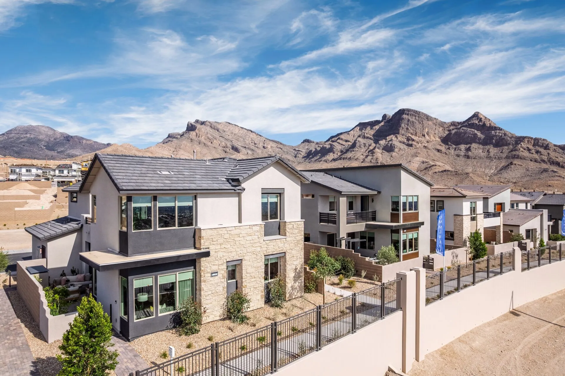 Modern two-story homes with stone and stucco exteriors sit on a landscaped street near desert mountains under a blue sky with wispy clouds. A metal fence lines the property, and sparse greenery accents the area.