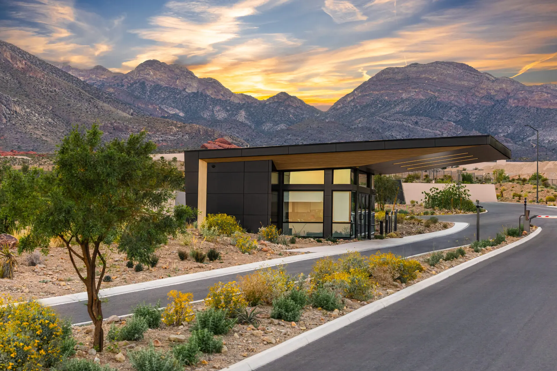 A modern building with large windows sits by a curved road, surrounded by desert plants and mountains under a dramatic, colorful sunset sky.