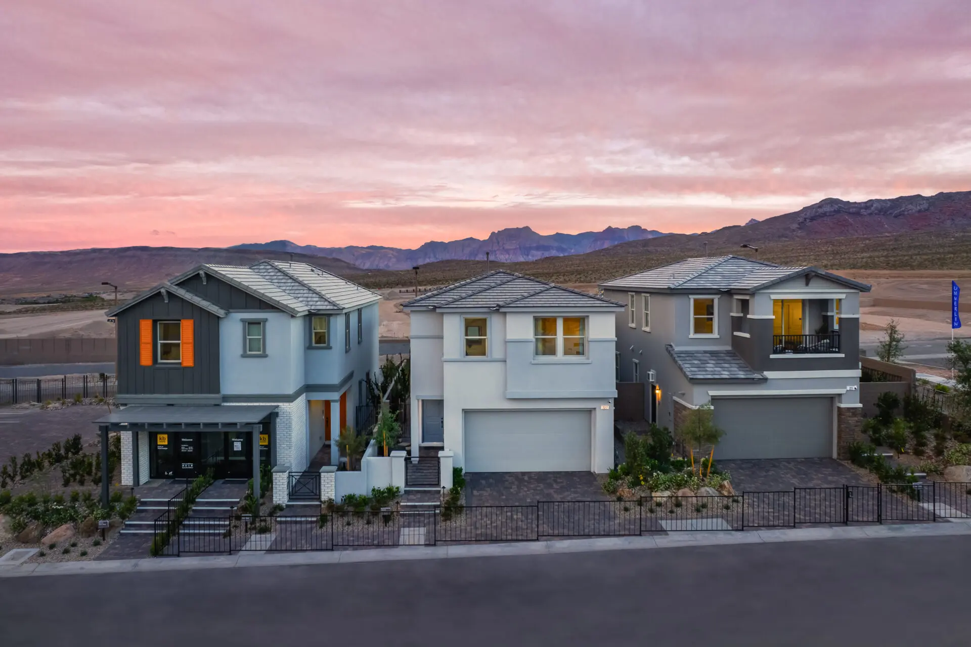 Three modern two-story houses with garages and landscaped yards are lined up on a street at sunset, with mountains and a colorful sky visible in the background.