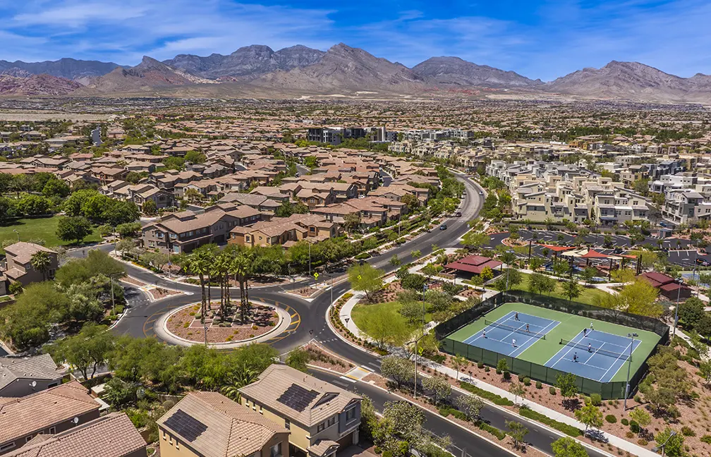 Aerial view of a suburban neighborhood with curved roads, rows of houses, tennis courts, palm trees, and mountains in the background under a blue sky.