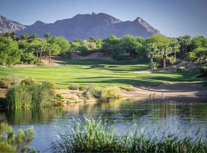Summerlin-Explore-Golf-Red-Rock-CC A lush golf course with a water hazard in the foreground, surrounded by trees and greenery, with tall mountains in the background under a clear blue sky.