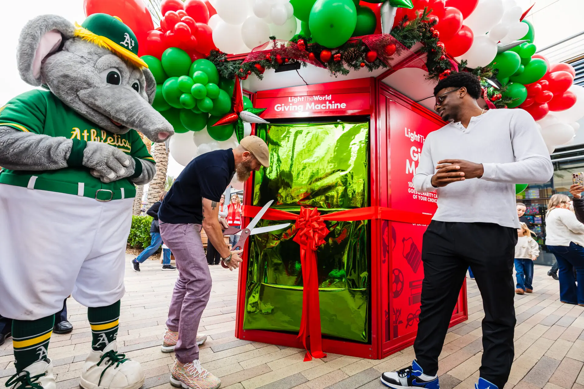 A man cuts a red ribbon on a large, gift-wrapped Giving Machine surrounded by balloons, as a mascot in an Oakland As uniform and others watch during an outdoor event.
