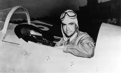Summerlin_About-Hughes-Communities A man wearing a flight suit, helmet, and goggles sits in the cockpit of an airplane, looking at the camera and smiling, in a black and white photograph.