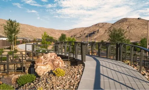 Summerlin_About-Maps-Guides A curved pedestrian bridge crosses a landscaped area with rocks, bushes, and trees, set against a backdrop of arid, brown hills under a partly cloudy sky.