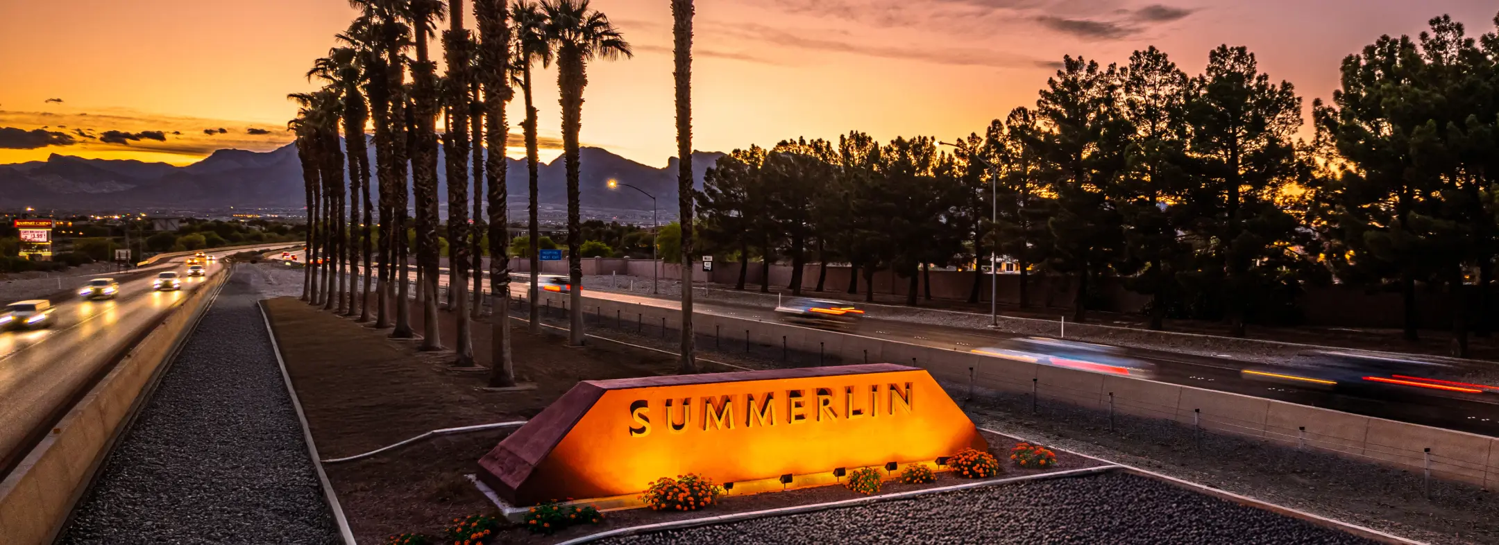 Summerlin_About A glowing Summerlin sign sits beside a road lined with palm trees at sunset, with cars passing by and mountains visible in the background.