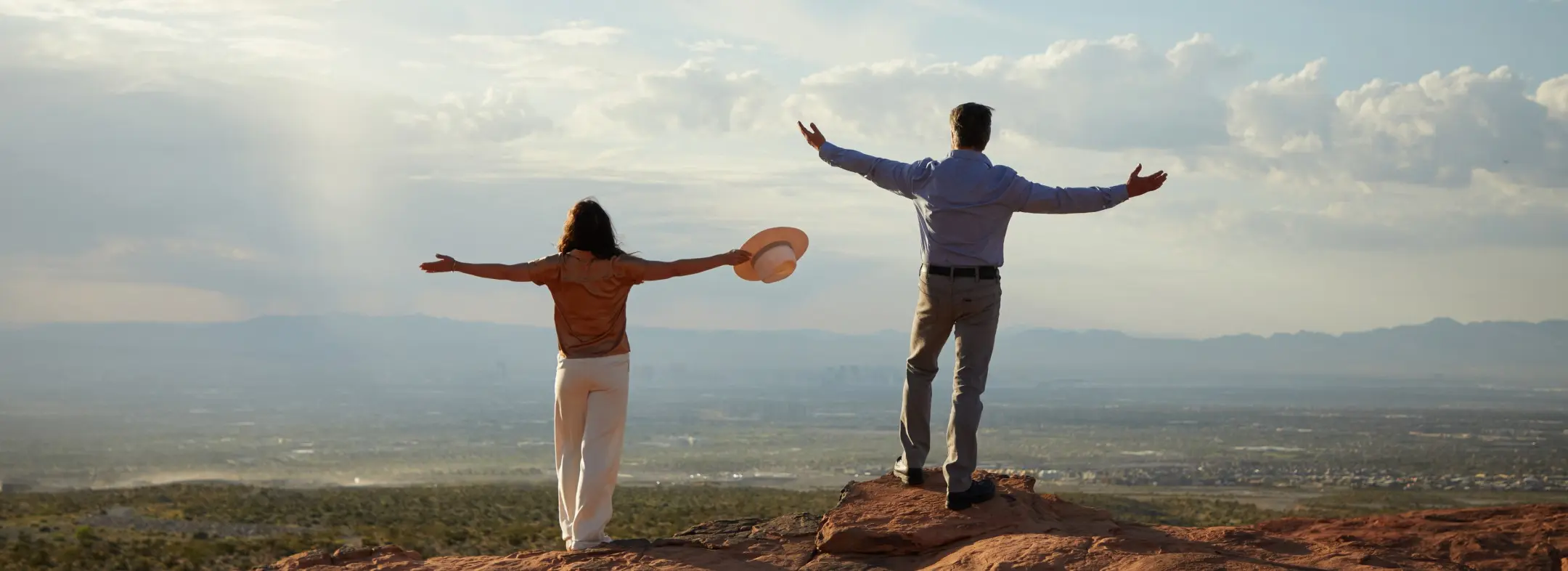 Summerlin_Custom-Astra Two people stand on a rocky hilltop with arms outstretched, looking out over a vast landscape under a partly cloudy sky. One holds a hat, and both appear to be enjoying the expansive view.