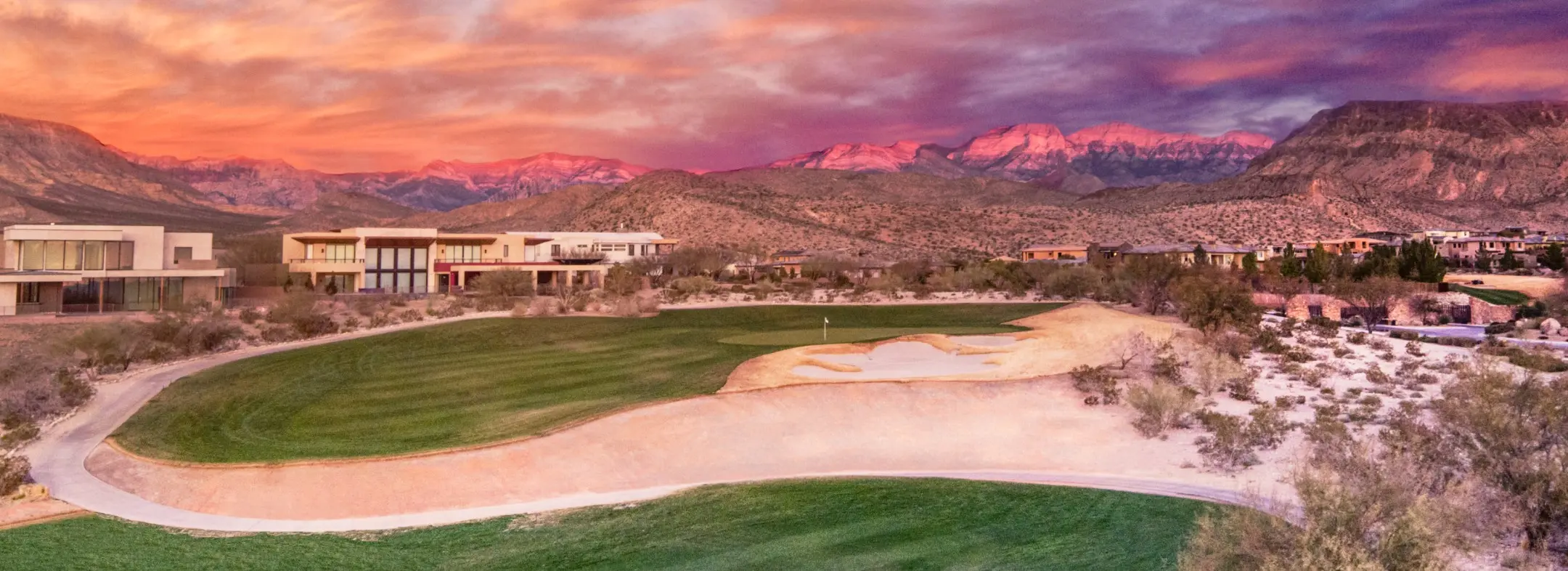 Summerlin_Custom-Ridges A golf course with sand bunkers and green grass is set against desert terrain and modern buildings. Purple and orange sunset clouds color the sky, and distant mountains glow pink in the background.