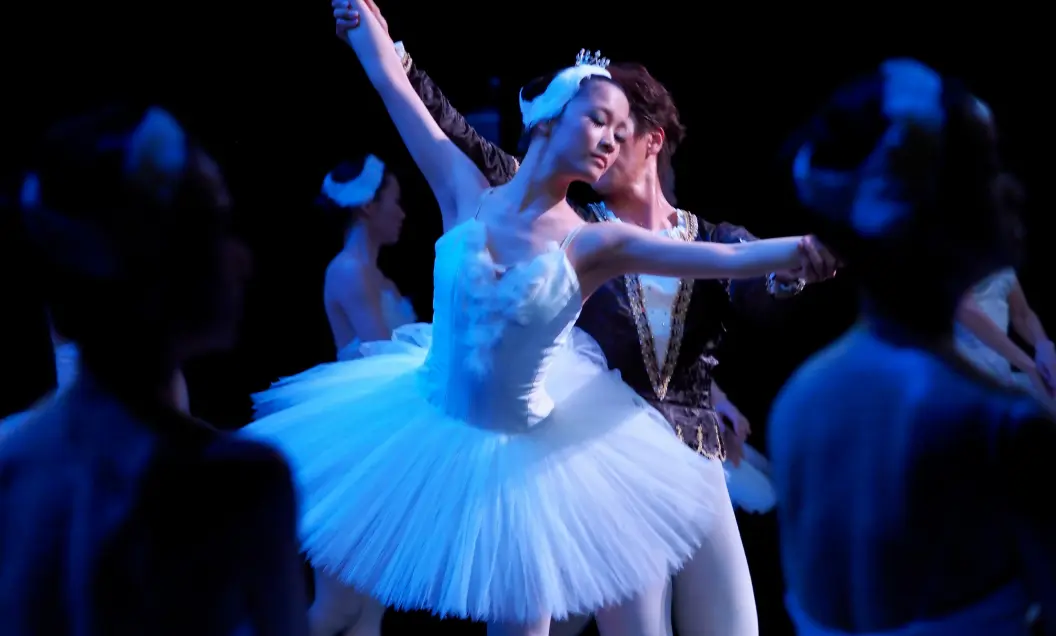 A ballerina in a white tutu and tiara dances gracefully with a male partner on a dimly lit stage, surrounded by other dancers in white costumes. The scene is illuminated with blue lighting.