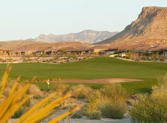 Summerlin_Explore-Golf-Amara A golf course with a flag on the green, surrounded by desert plants, houses in the background, and rocky mountains under a clear sky.