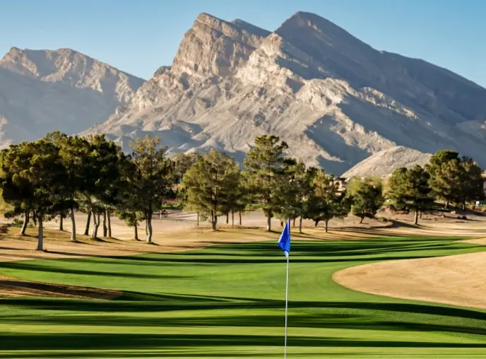 Summerlin_Explore-Golf-Eagle-Crest A green golf course with a blue flag on the putting green, surrounded by trees and dramatic mountain peaks in the background under a clear blue sky.