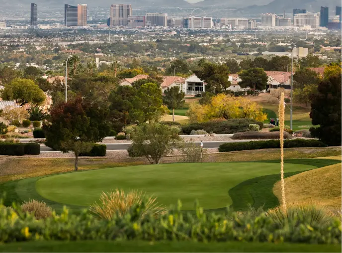 Summerlin_Explore-Golf-Highland-Falls A lush green golf course with trees and shrubs in the foreground, suburban houses in the middle, and a city skyline with tall buildings and mountains in the background.
