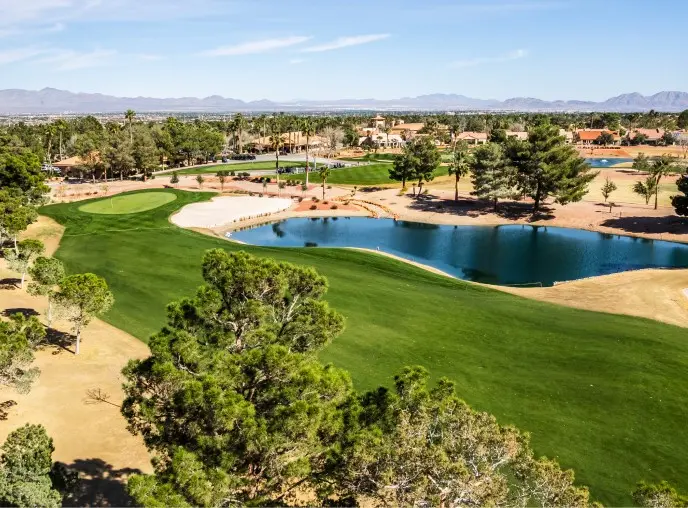 Summerlin_Explore-Golf-Palm-Valley-GC A lush golf course with green fairways, sand bunkers, a pond, and trees, surrounded by houses and distant mountains under a clear blue sky.