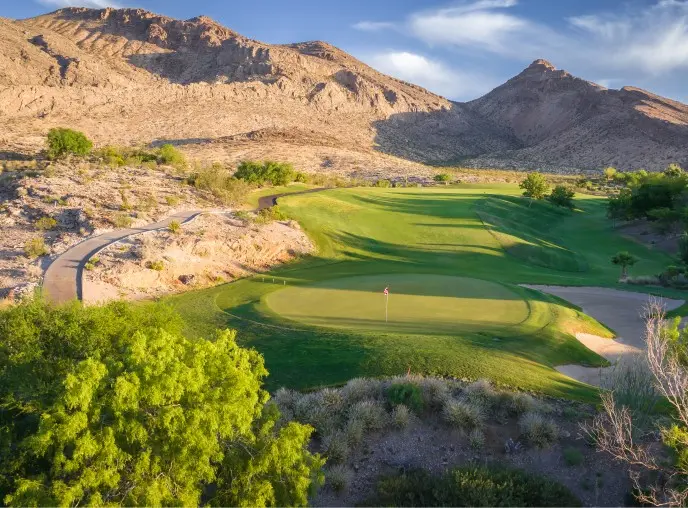 Summerlin_Explore-Golf-Red-Rock A lush green golf course sits amid desert terrain and rugged mountains under a blue sky with wispy clouds, blending natural scenery with manicured landscape.