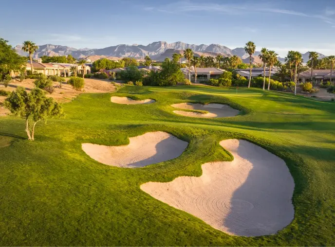 Summerlin_Explore-Golf-Siena_GC A lush golf course with several sand bunkers, palm trees, and nearby houses, set against a backdrop of distant mountains under a clear blue sky.