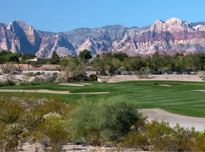Summerlin_Explore-Golf-TPC-Las-Vegas A lush green golf course sits in the foreground, bordered by desert shrubs and trees, with red and tan rocky mountains rising dramatically in the background under a clear blue sky.