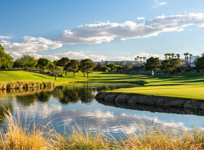 Summerlin_Explore-Golf-TPC_Summerlin A scenic golf course with lush green fairways, trees, and palm trees is reflected in a calm water hazard under a blue sky with scattered clouds. Dry grasses frame the foreground.