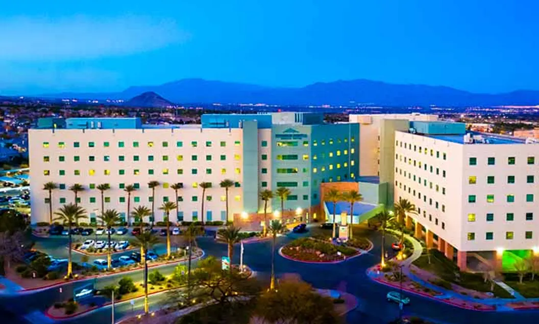 An aerial view of a large, modern hospital complex at dusk, with illuminated windows, palm trees, roundabout drives, and a cityscape and mountains visible in the background.