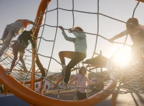 Children climb and play on a large orange rope climbing structure at a playground, with the sun shining brightly in the background.