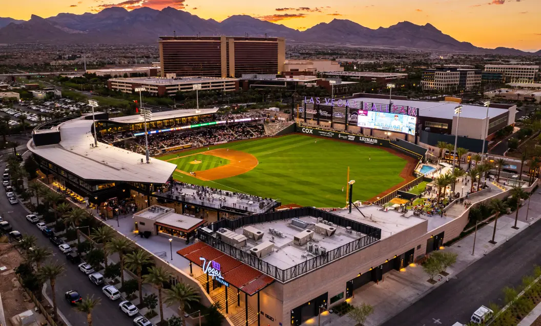 Aerial view of a baseball stadium at sunset, surrounded by palm trees, city buildings, and parking lots, with mountains visible in the background. The field lights are on and the scoreboard is illuminated.