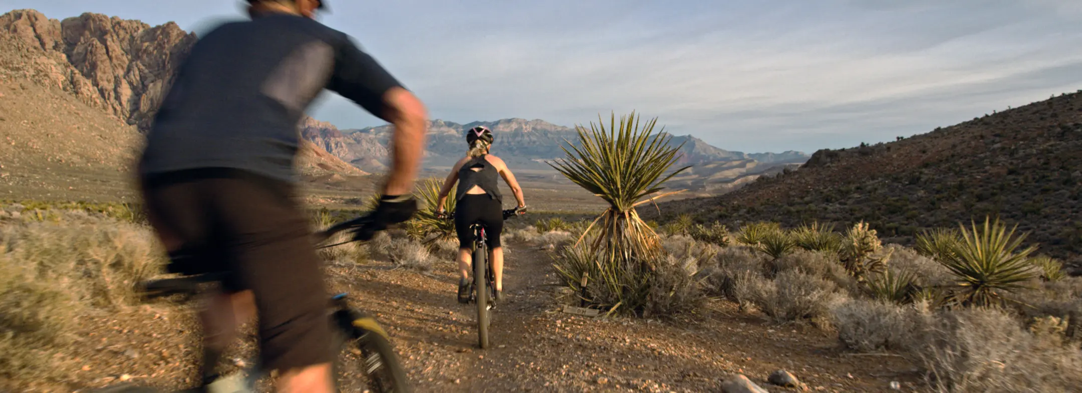 Two people ride mountain bikes on a rocky desert trail surrounded by shrubs and yucca plants, with distant mountains under a partly cloudy sky. The scene is sunlit and outdoorsy.