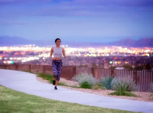 A woman runs on a paved path in a park at dusk, with city lights glowing in the blurred background and mountains in the distance. She wears a white tank top and colorful leggings.