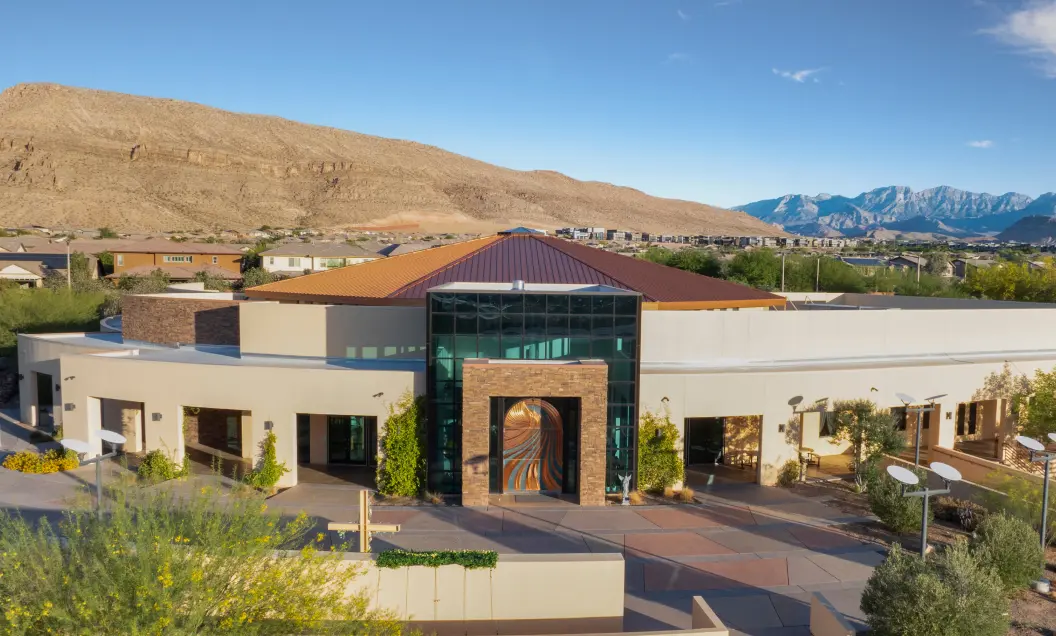 A modern building with a circular design, large windows, and a decorative entrance sits amid desert landscaping, with mountains and a clear blue sky in the background.
