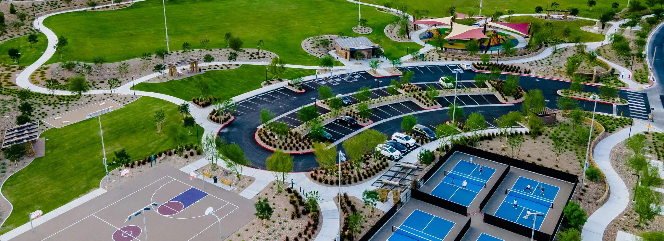Summerlin_Parks-Amenities Aerial view of a park with green lawns, walking paths, a basketball court, multiple pickleball courts, a shaded playground area, and a parking lot with several cars. Trees and landscaping are spread throughout the park.