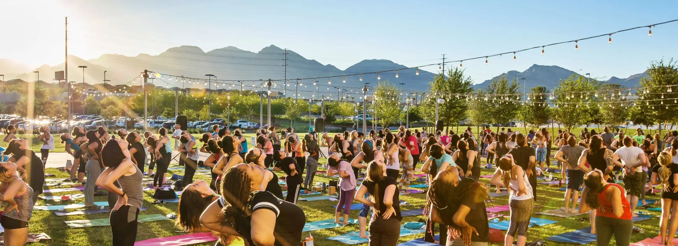 Summerlin_Parks-Community A large group of people practice yoga outdoors on a grassy field at sunset, surrounded by trees and mountains, with string lights hanging overhead.