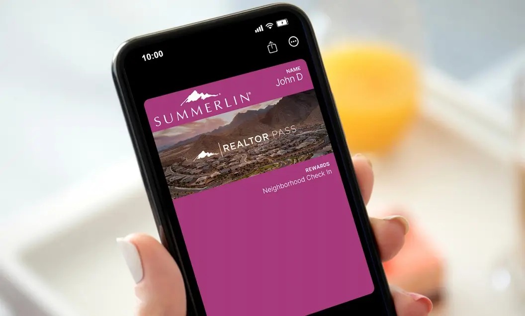 A hand holds a smartphone displaying a digital Summerlin Realtor Pass for John D with a mountain logo, scenic photo, and Neighborhood Check In rewards, against a blurred background.
