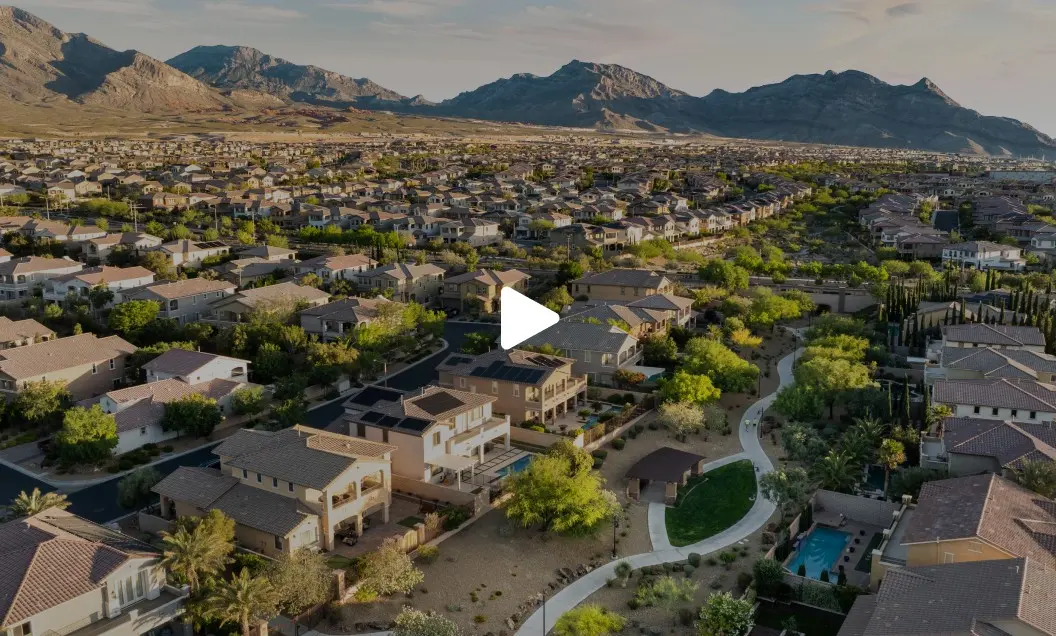 Aerial view of a suburban neighborhood with rows of houses, green trees, and a winding walking path, set against a backdrop of mountains under a clear sky.
