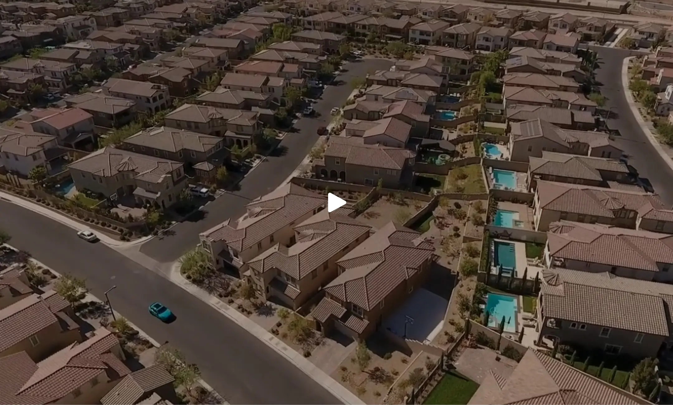 Summerlin_Sustainability-Neighborhood-Video-Placeholder Aerial view of a suburban neighborhood with rows of similar houses, some with blue swimming pools in their backyards. A single blue car is driving along a road in the lower left corner.