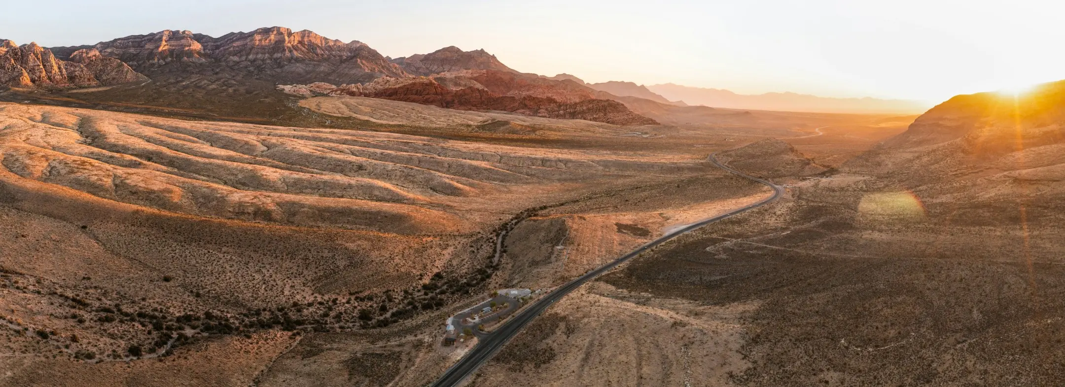 Summerlin_Sustainability-Red-Rock-Canyon A panoramic view of a desert landscape at sunset, featuring a winding road, rugged mountains in the background, and golden sunlight illuminating the dry, rolling terrain.